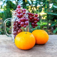 two oranges and a glass jar with grapes on a wooden tabletop against a background of green vegetation